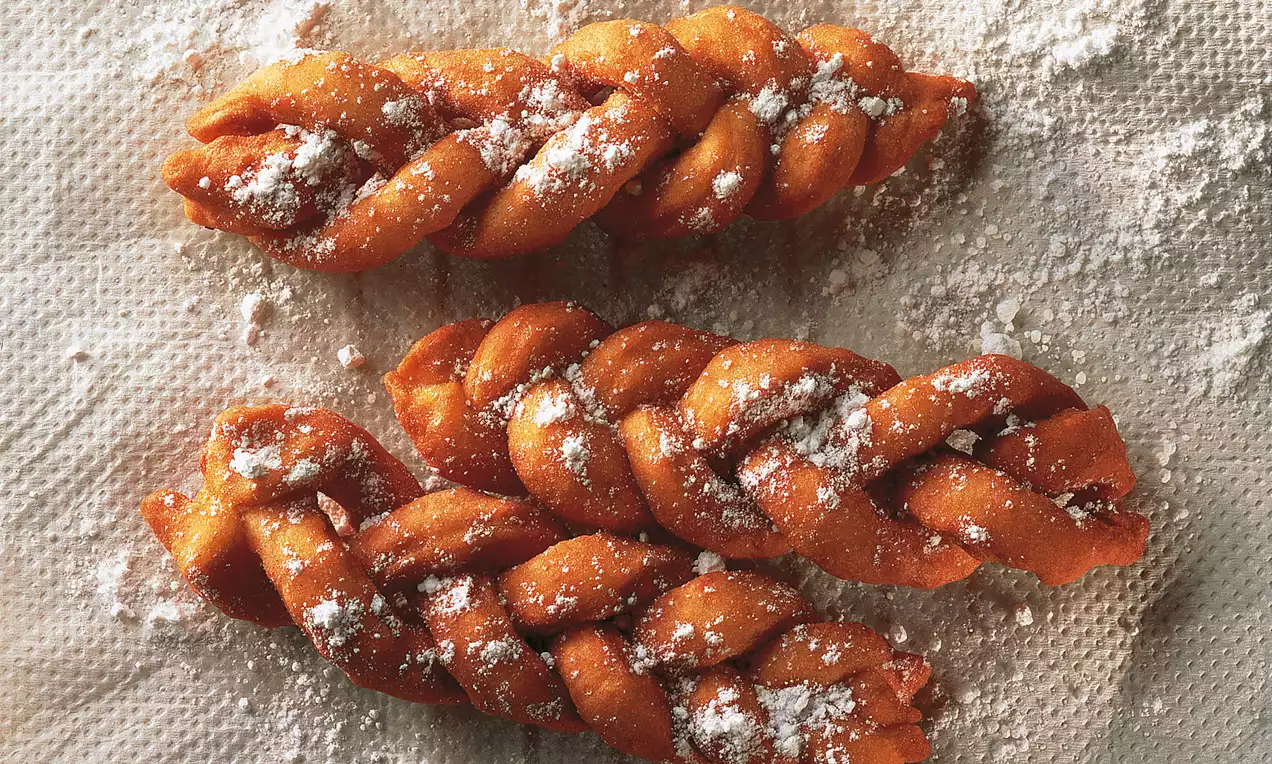 Drei goldbraune, geflochtene Fasnachtszöpfchen mit knuspriger Textur, leicht mit Puderzucker bestäubt auf einem weiß strukturierten Untergrund