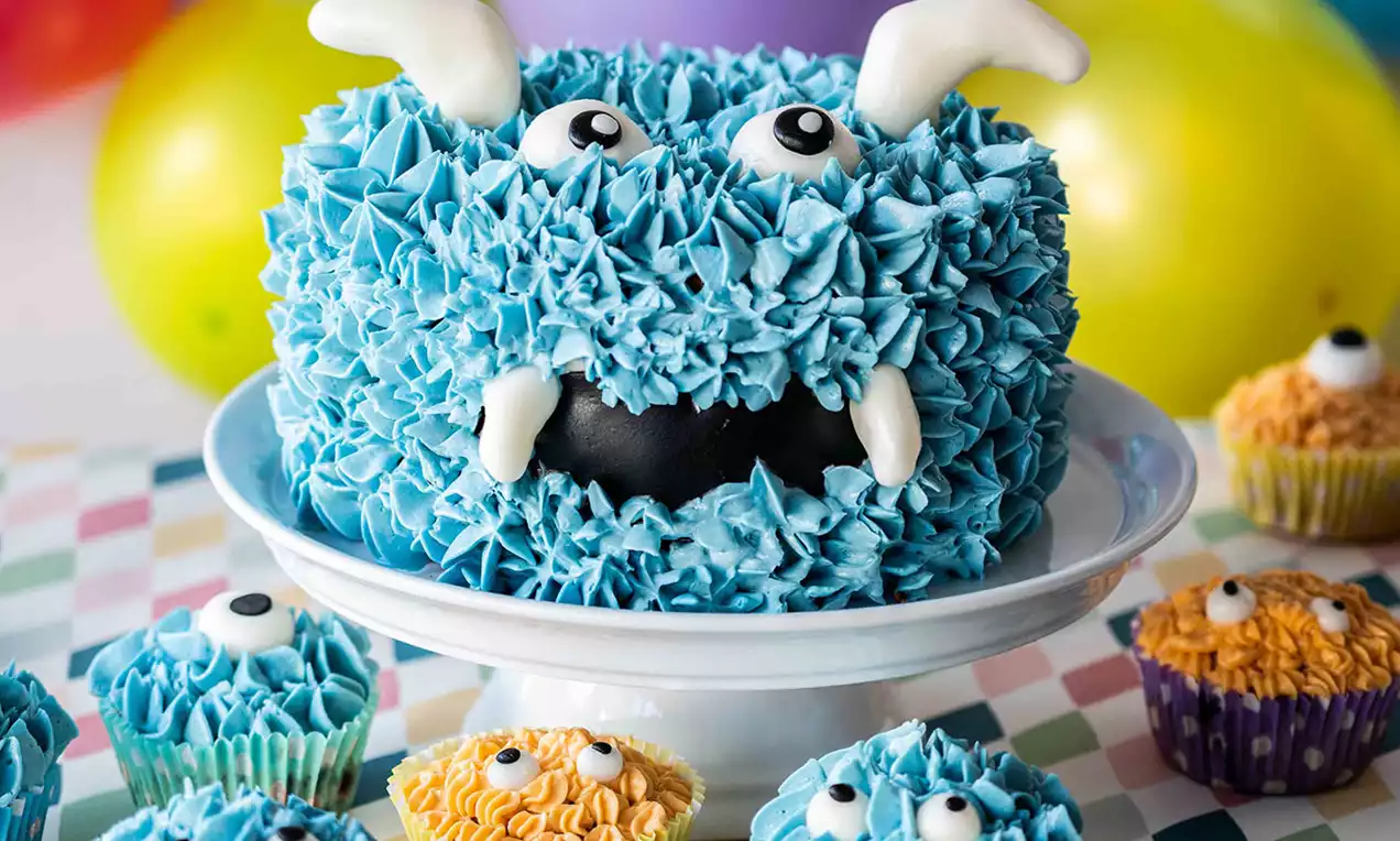 Runde Torte mit blauer Creme als flauschiges Fell, weißen Zähnen, großen Augen und Hörnern auf einer weißen Tortenplatte mit bunten Mini-Cupcakes