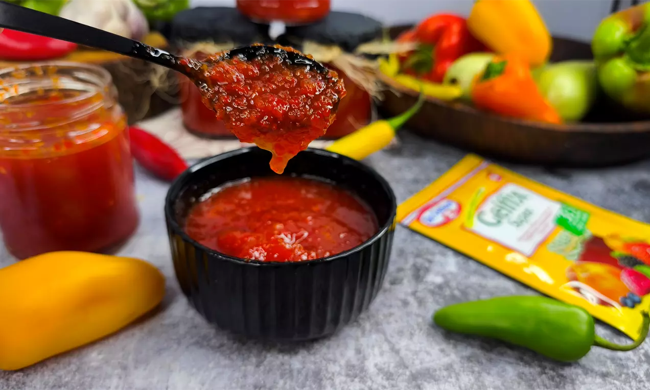 Гъст червен доматен сос с текстура на ситно нарязани парчета в черна купа върху сив плот, заобиколена от пресни разноцветни чушки