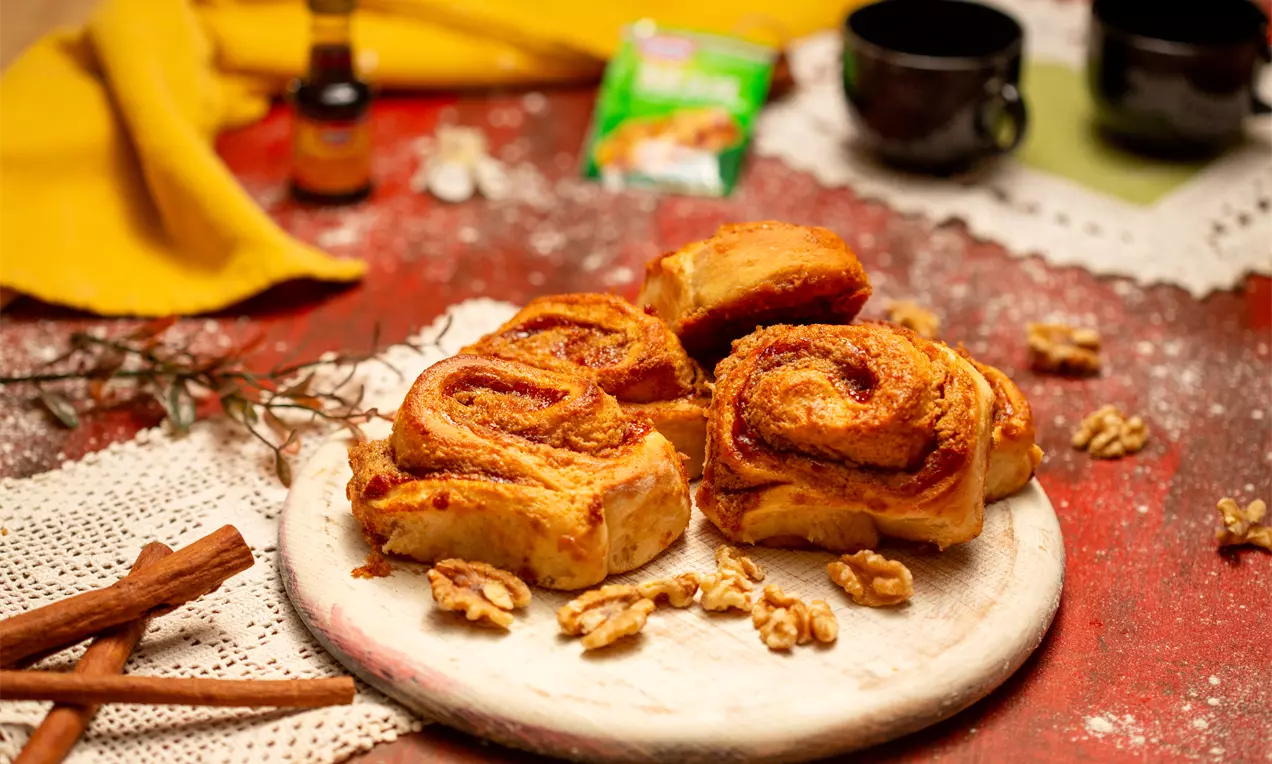 Златисто-кафяви козуначени рула с рехава текстура, наситени с орехи и мармалад, подредени върху бяла дървена дъска с разпилени орехи