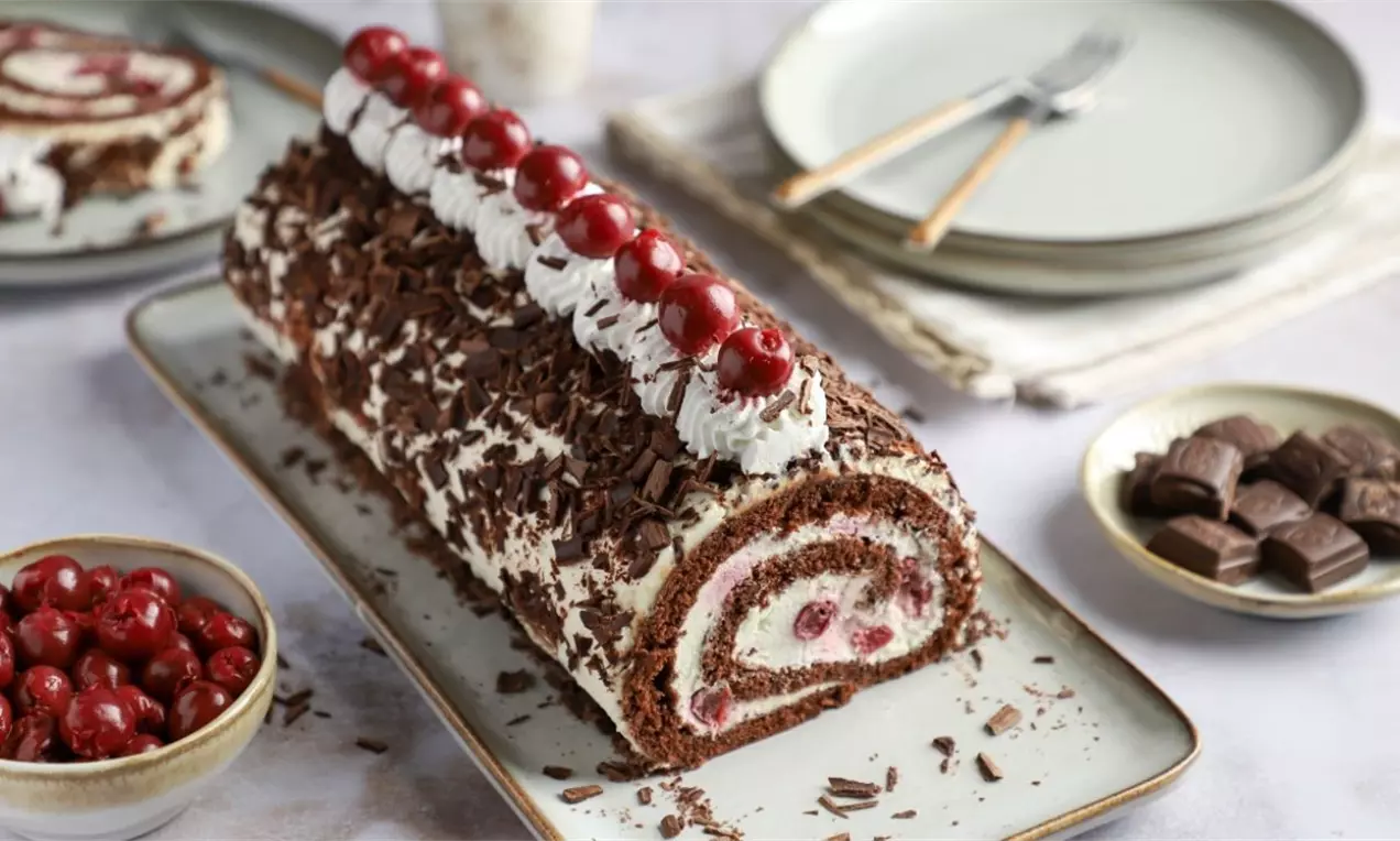 Tamnosmeđi čokoladni rolat ukrašen šlagom, crvenim trešnjama i čokoladnim strugotinama, serviran na tanjiru uz posudu s trešnjama