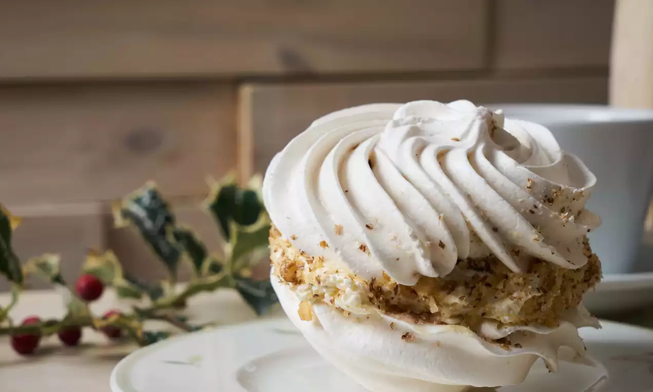 Bezea albă crocantă, umplută cu cremă bogată și decorată cu firimituri aurii de nucă, așezată pe o farfurie lângă decorațiuni festive verzi și roșii