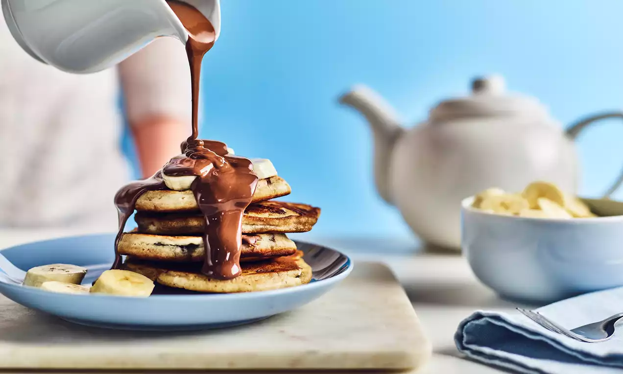 A stack of golden-brown gluten-free pancakes with chocolate sauce poured over, topped with banana slices on a blue plate