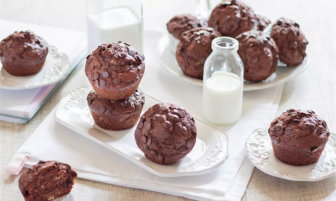 Шоколадови мъфини с наситен кафяв цвят, кръгла форма и влажна текстура, поръсени с шоколадови капки, сервирани върху бели чинии