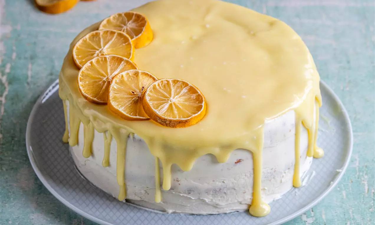 Кръгла лимонова торта с крем маскарпоне, покрита с жълт глазур с капки по краищата и декорирана с тънки резени лимон върху светла чиния