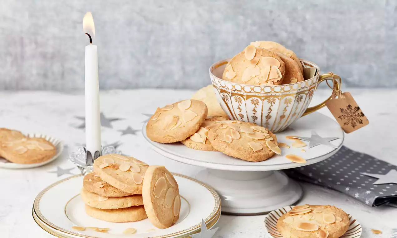 Goldbraune Mandelkekse mit knusprigen Mandelblättchen auf edlen weißen Tellern und einer verzierten Tasse, dekoriert mit silbernen Sternen