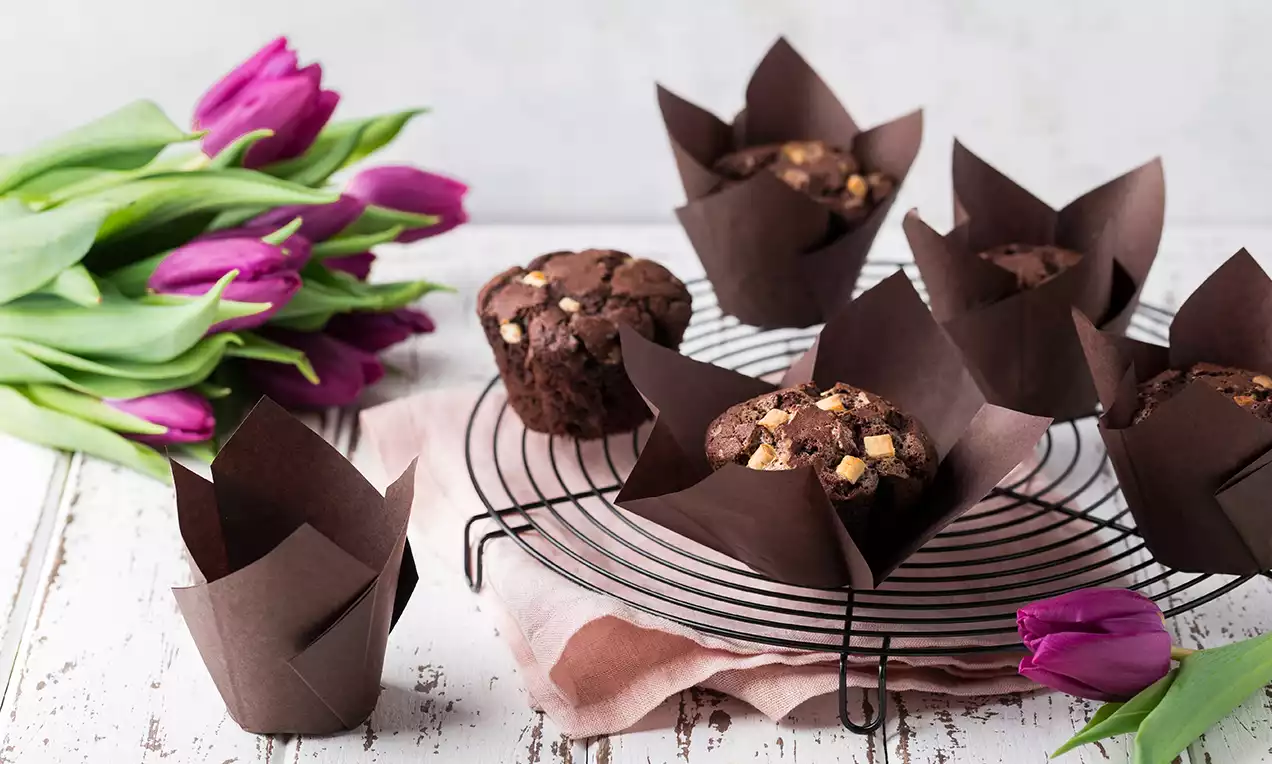 Muffins au chocolat foncé enveloppés dans des caissettes tulipes marron sur une grille noire, accompagnés de tulipes violettes sur une table blanche