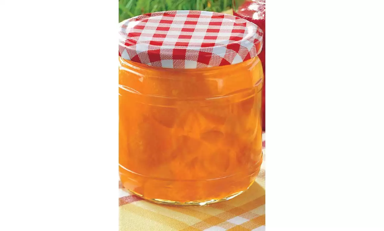 Glazen pot gevuld met feloranje abrikozenjam, afgesloten met een wit-rood geruit deksel, rustend op een geel tafelkleed