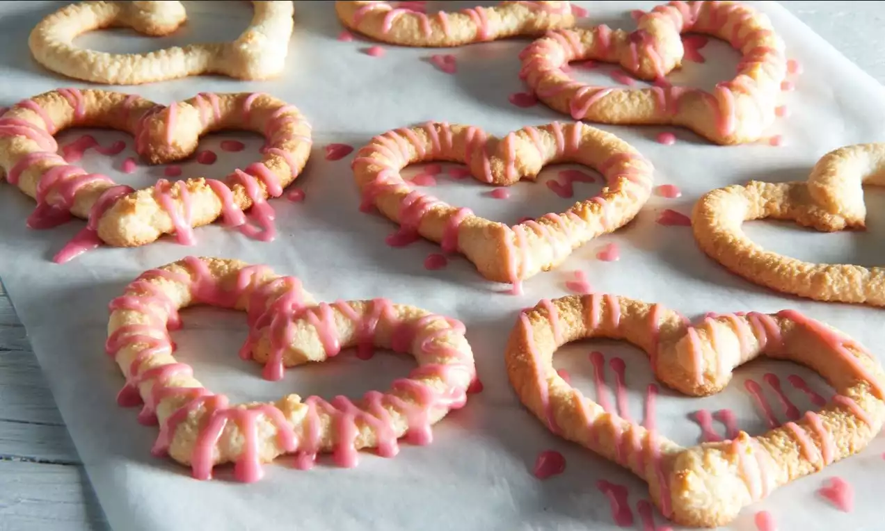 Marcipánová srdíčka světle zlatavá, měkké textury, zdobená růžovou polevou, rozmístěná na pergamenovém papíru v nepravidelném uspořádání