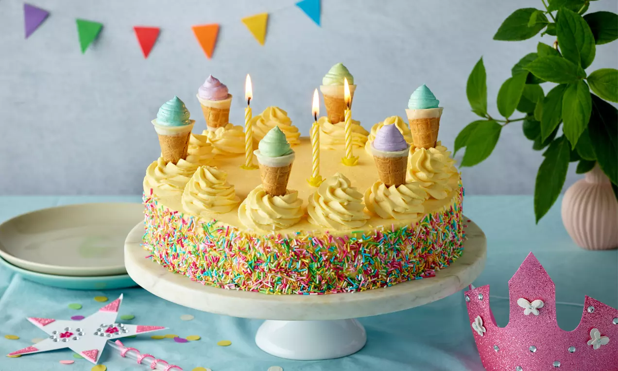 Runde Geburtstagstorte mit gelber Creme, bunten Streuseln und kleinen Eiswaffeln, darauf Kerzen, auf weißer Tortenplatte arrangiert