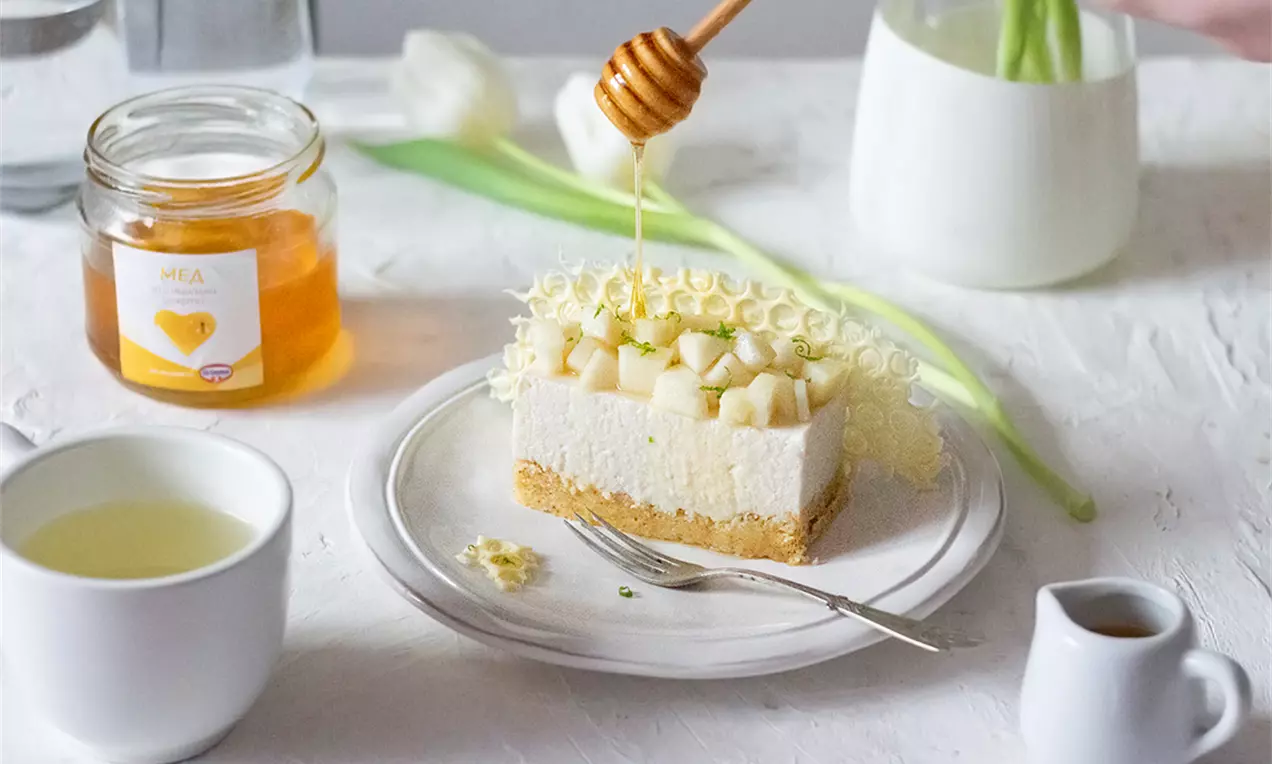 Кремообразен чийзкейк с меден вкус върху златист бисквитен блат, гарниран с кубчета сочна круша и къдрици бял шоколад върху бяла чиния