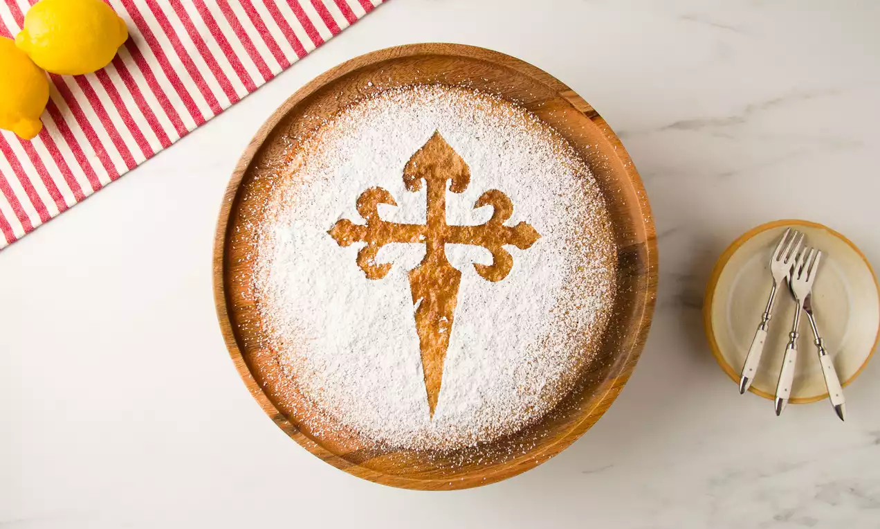 Tarta redonda de almendra dorada con superficie espolvoreada de azúcar glas formando la cruz de Santiago, sobre un plato de madera