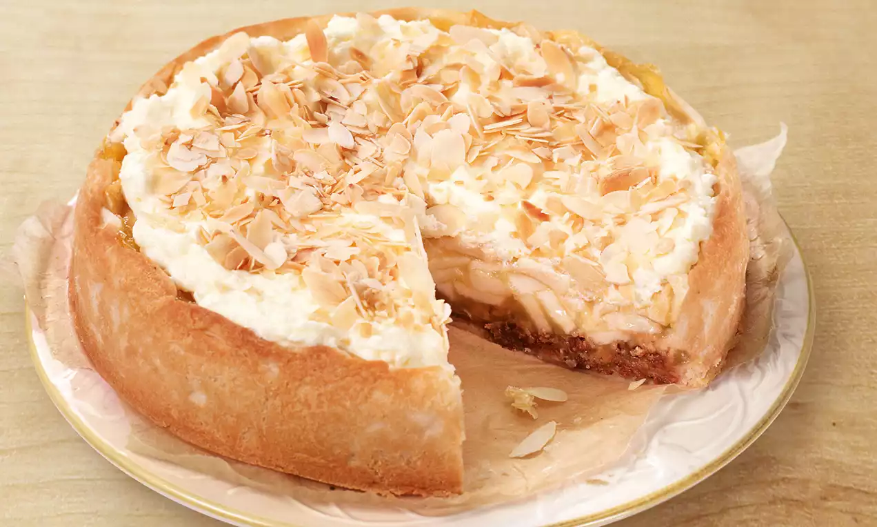 Torta dorata con base croccante, ripieno cremoso sormontato da mele sottili e fiocchi di mandorle su un piatto bianco ornato