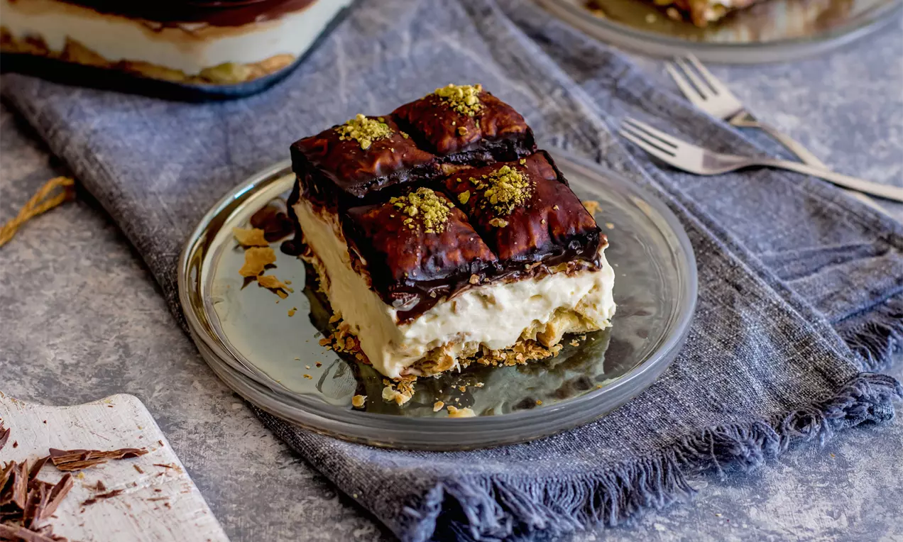 Квадратен десерт Наполеон с блестяща шоколадова глазура, поръсена с шамфъстък, върху мек крем и хрупкаво маслено тесто в стъклена чиния