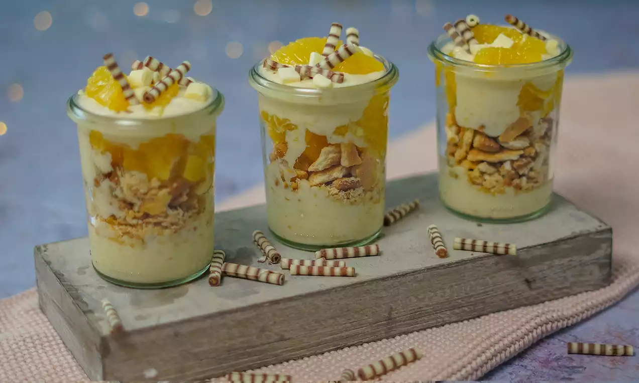 Dessert étagé en verrine avec crème jaune pâle, morceaux d’orange, biscuit émietté, décoré de rouleaux chocolatés sur une planche