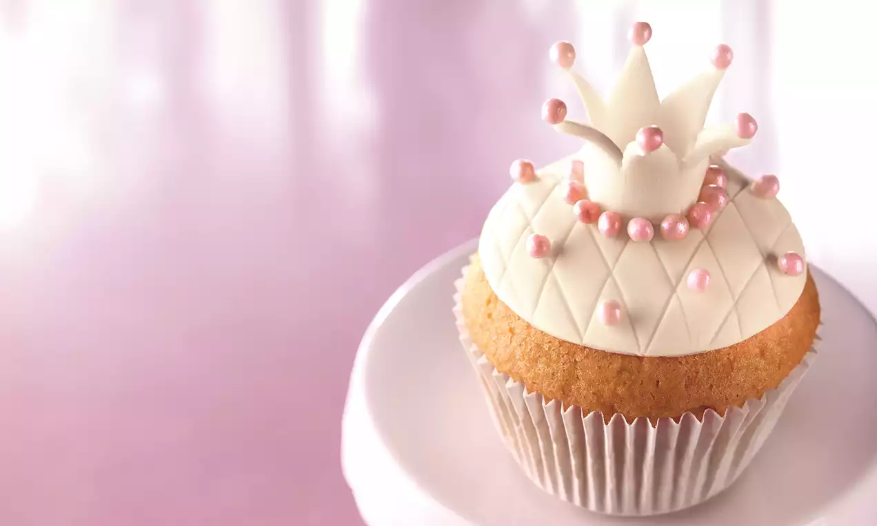Babeczka z jasnego ciasta w białej papilotce, zwieńczona kremem w kształcie korony, dekorowana perełkami w odcieniach różu i bieli