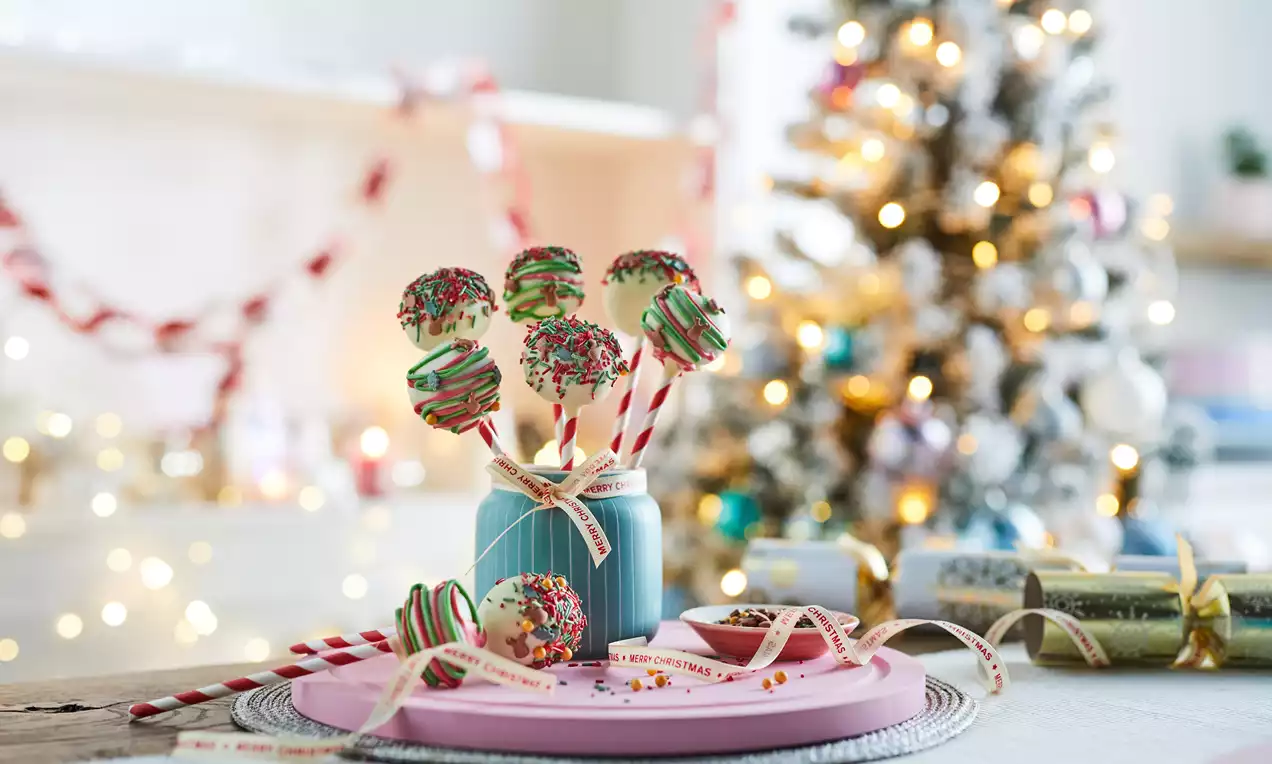 Festive cake pops in rich green and red swirls with sprinkles on striped sticks, arranged in a jar and on a pink plate amidst Christmas decor
