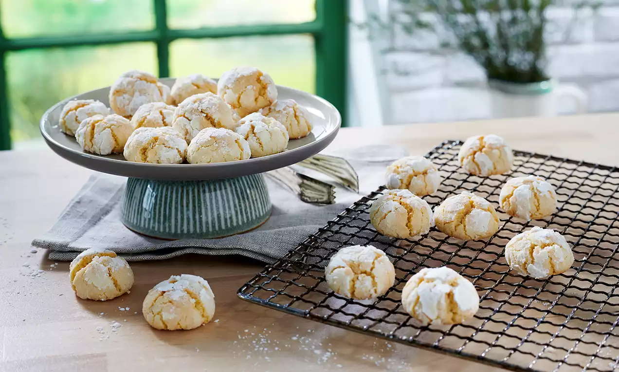 Svijetložuti raspucanci s bijelim šećerom u prahu, okrugli i neravne površine, raspoređeni na žičanom stalku i plavoj keramičkoj zdjeli