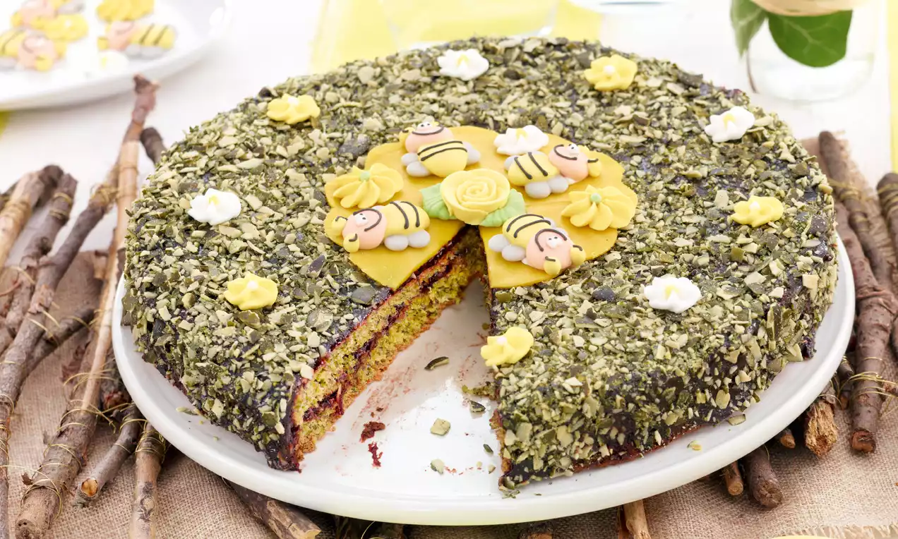 Runder Kürbiskernkuchen mit grünem Kernmantel, bunter Schichtfüllung und Deko aus gelben Marzipanbienen und Blüten auf weißem Teller