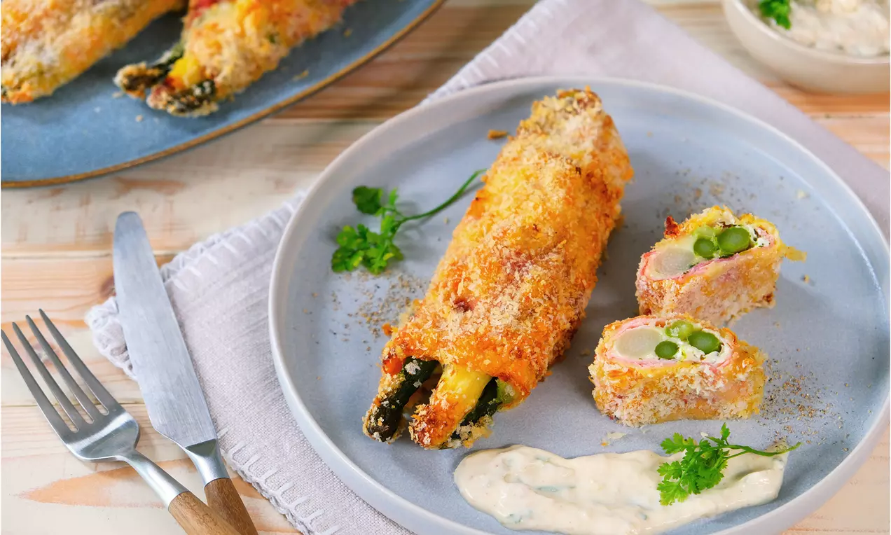 Goldbraun paniertes Spargel Cordon Bleu mit knuspriger Kruste, grünem Spargelkern und Beilage aus cremiger Sauce auf grauem Teller
