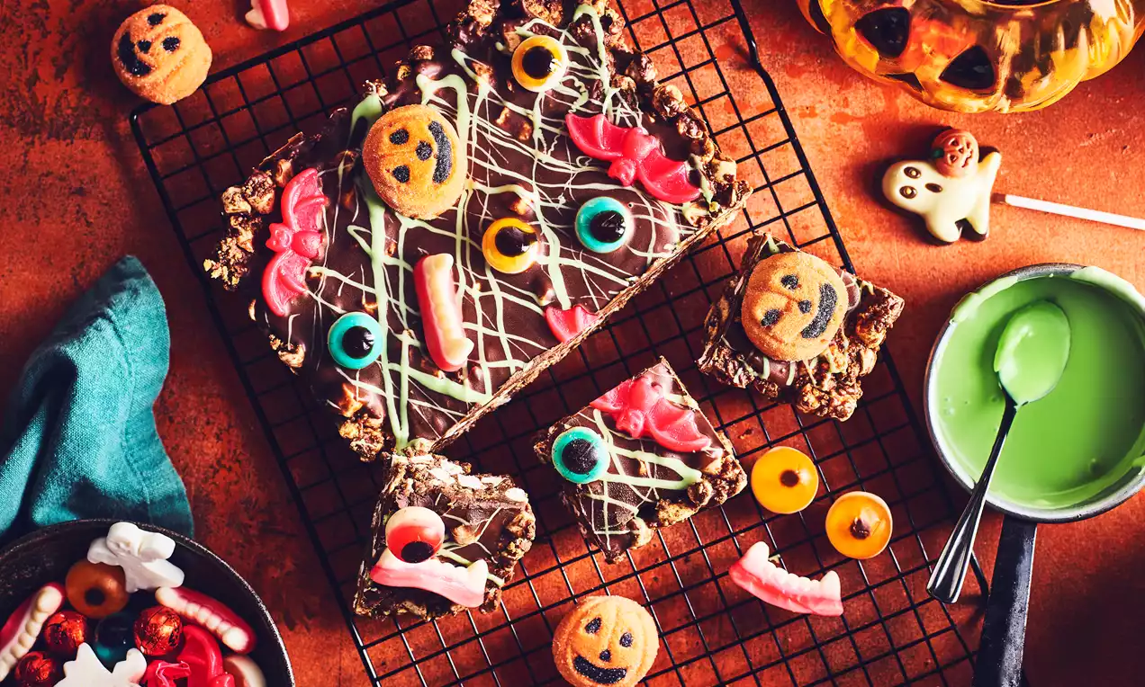 Rocky road squares topped with drizzled green and white chocolate, gummy candies, and smiling orange pumpkin biscuits on a black wire rack