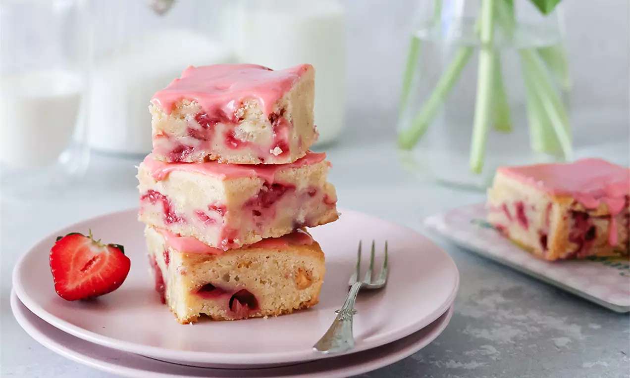 Бледорозови квадратчета с кремообразна текстура, поръсени с ягодови парченца, наслояват се върху розова чиния до разрязана ягода