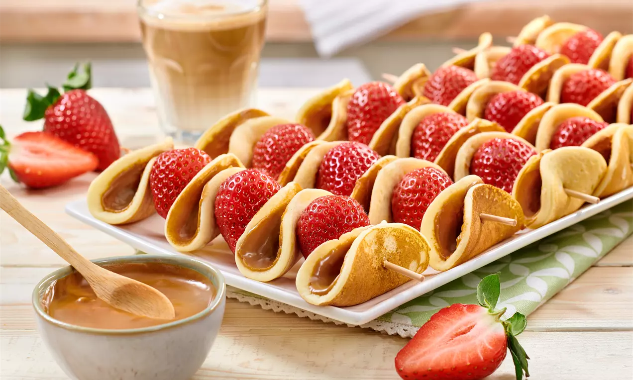 Brochettes alignées de mini crêpes dorées pliées autour de fraises rouges brillantes sur un plat rectangulaire avec une sauce caramel