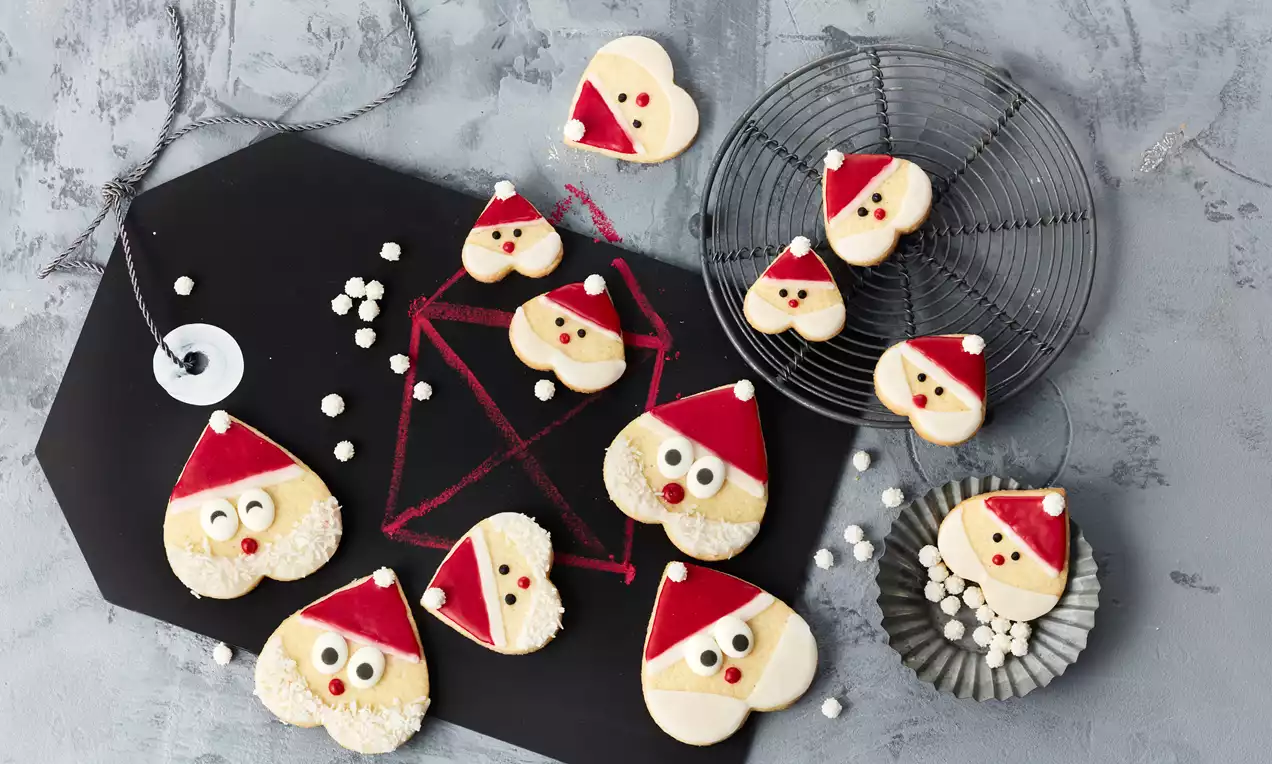 Herzförmige Kekse mit roter Zuckergussmütze, weißen Augen und cremefarbenem Bart auf schwarzem Holzbrett und silbernem Gitter arrangiert