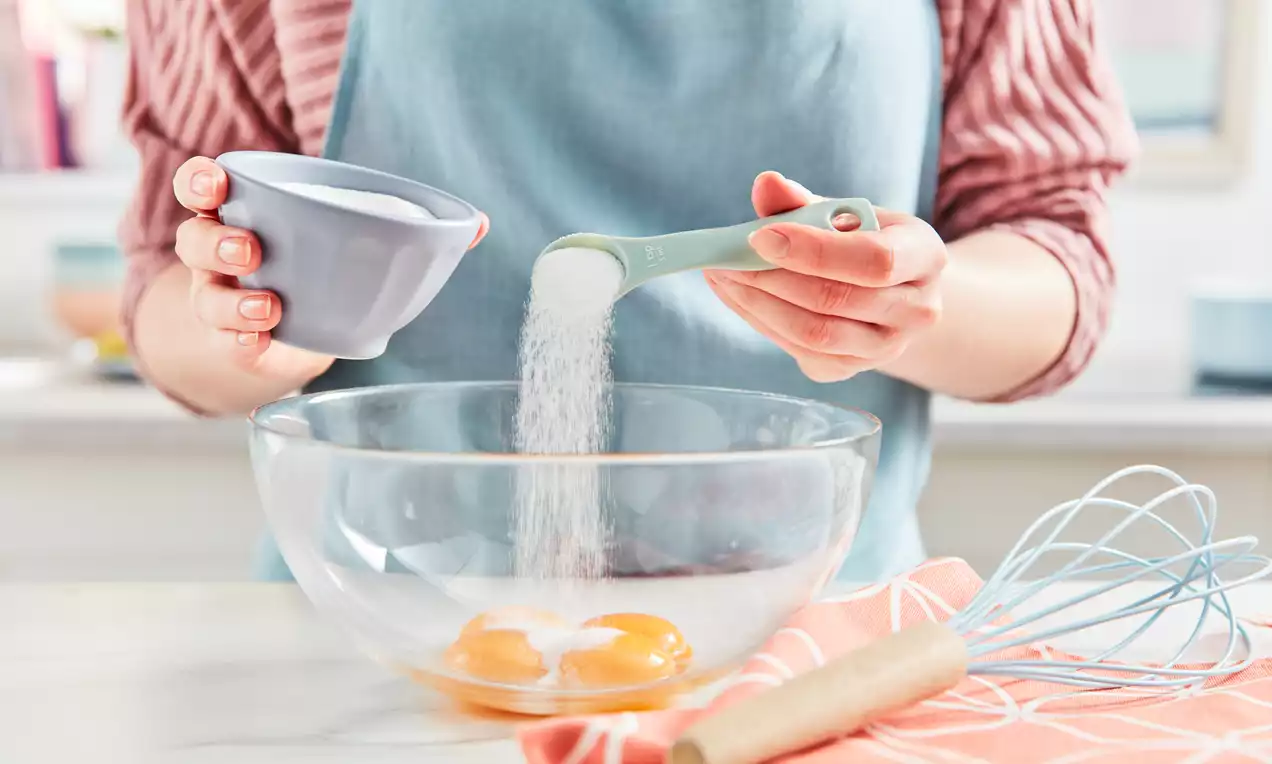 Two golden egg yolks in a clear glass bowl on a white countertop, with sugar being poured from a pastel green spoon by a person in a blue apron