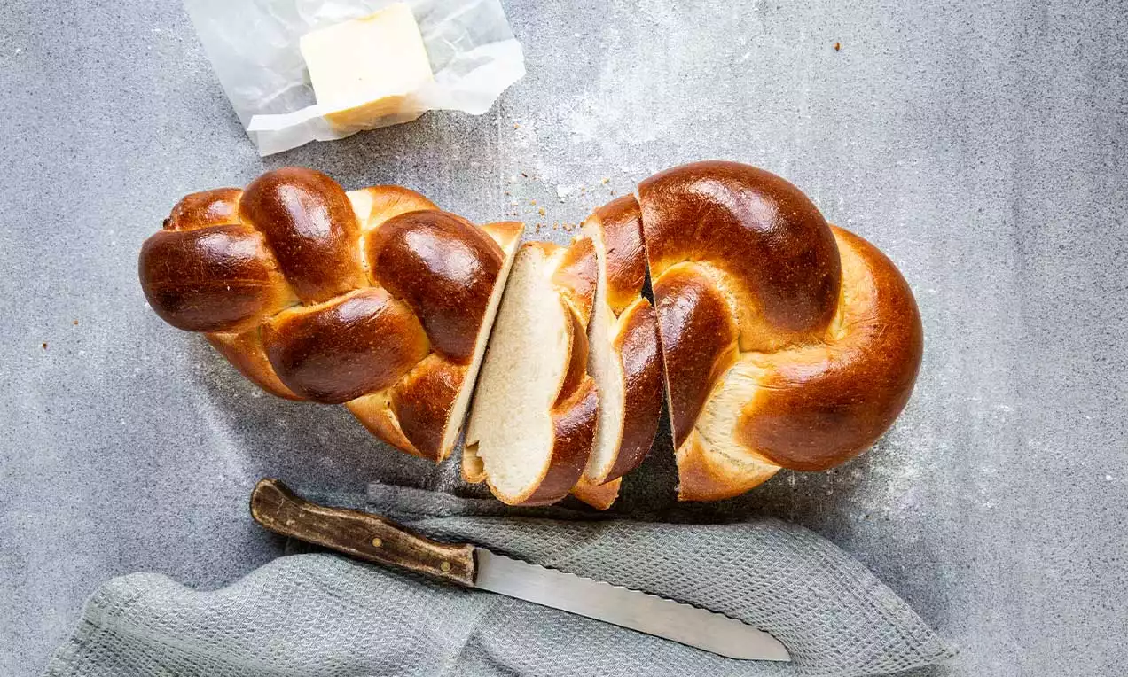 Butterzopf mit goldbrauner glänzender Kruste, geflochtene Form, teilweise aufgeschnitten, auf grauer Oberfläche mit Brotmesser und Butterstück