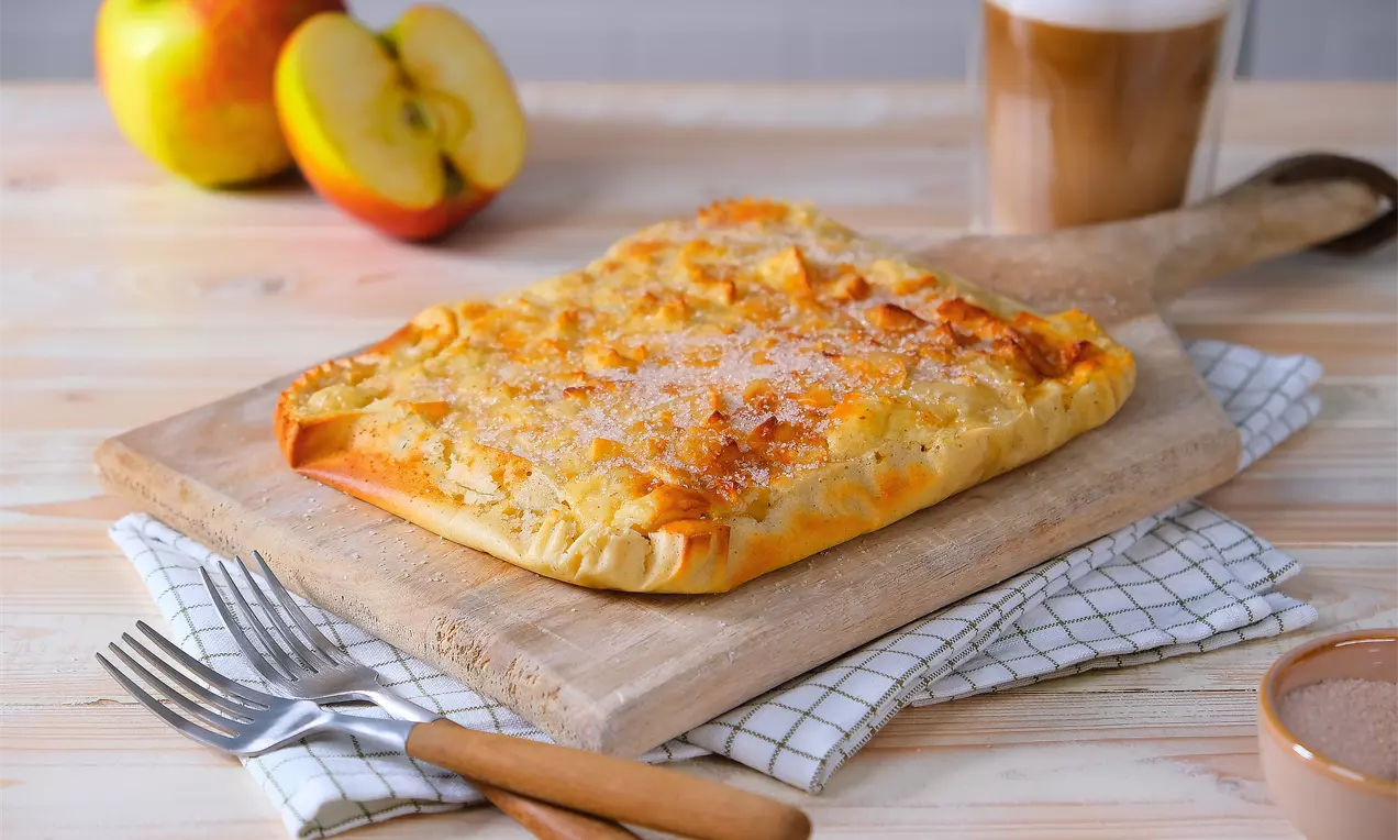 Rechteckiger, goldbraun gebackener Pfannkuchen mit feiner Zuckerkruste auf einem Holzbrett, umgeben von Äpfeln und Getränk