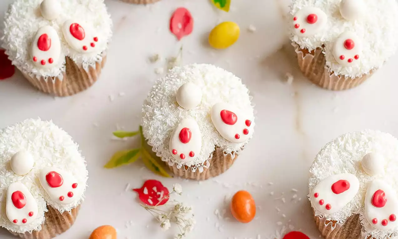 Cupcakes s mekanim tijestom u smeđim košaricama, prekriveni bijelim kokosom i dekorirani kremom u obliku zečjih šapica na bijeloj podlozi s cvjetnim ukrasima