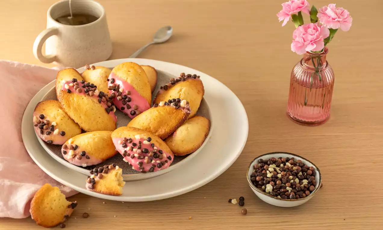 Madeleines dorées et moelleuses, partiellement enrobées de glaçage rose et parsemées de billes chocolatées sur une assiette blanche