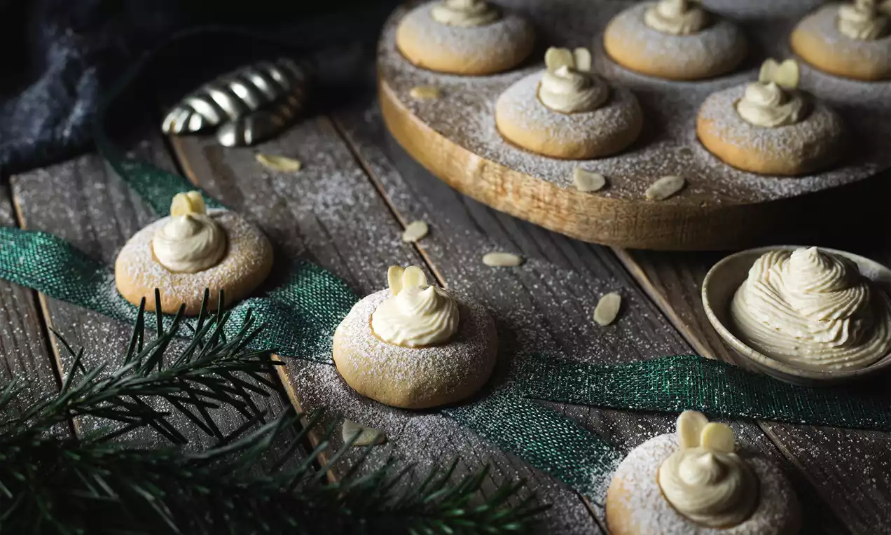 Kulaté máslové sušenky se světlým likérovým krémem, posypané moučkovým cukrem a zdobené plátky mandlí na dřevěném stole