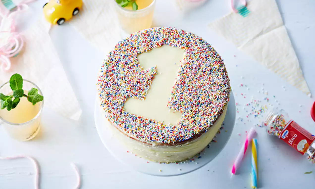 Gâteau rond à base crème claire avec le chiffre 1 en glaçage vanille, entouré de vermicelles colorés sur une assiette blanche