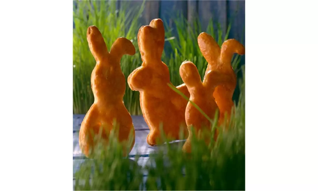 Petits lapins dorés en pâte levée, aux oreilles dressées, disposés debout au milieu de brins d'herbe verte sur une table en bois