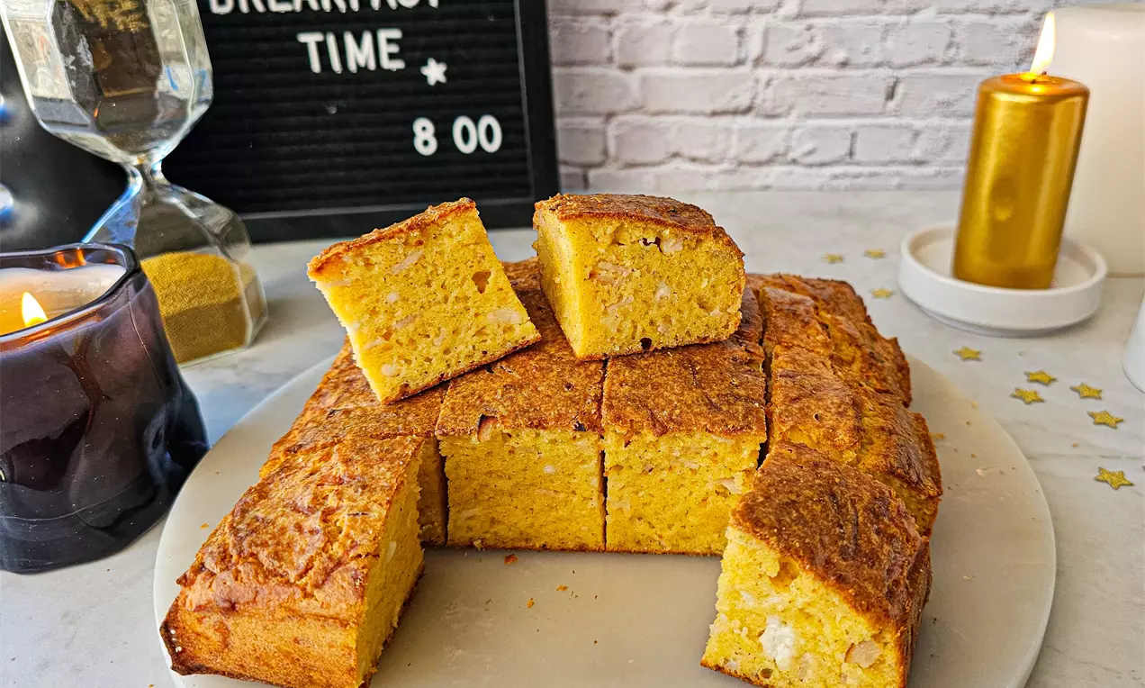 Златисто-кафяв солен кекс с неравномерна текстура, парченца сирене и шунка, нарязан на кубчета върху бяла чинийка