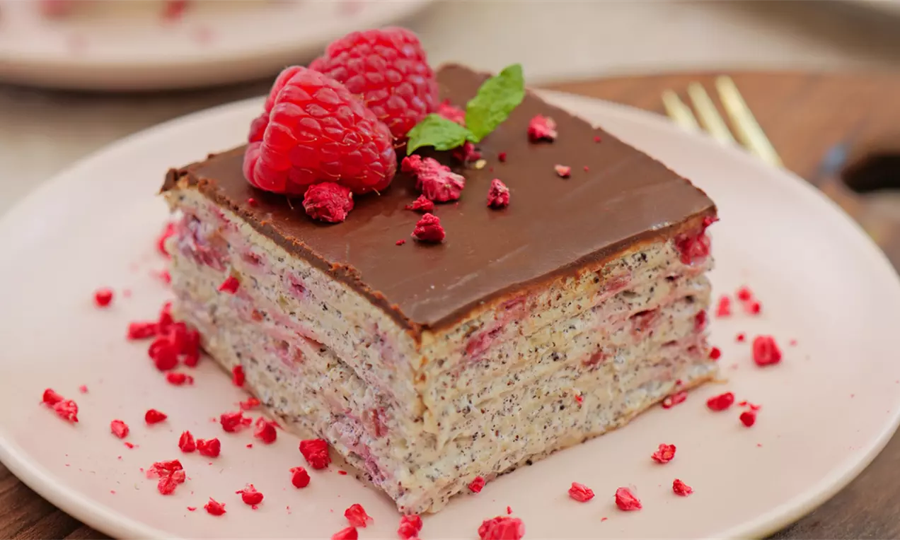 Kockasta torta sa slojevima svetle mak kore i roze fila od malina, prekrivena glatkim čokoladnim prelivom, ukrašena svežim malinama i listićem mente