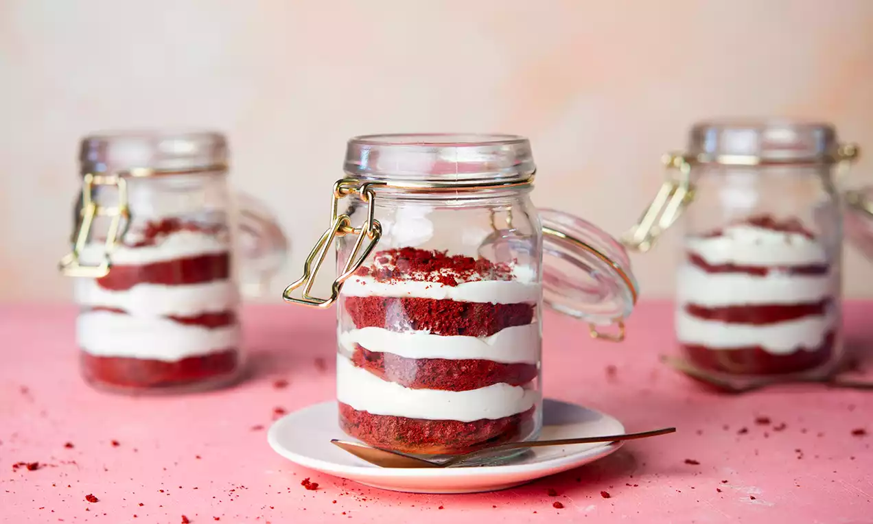Gelaagde trifle in een glazen potje met diepe rode cake en witte roomlagen, gepresenteerd op een klein wit bord met een lepel ernaast