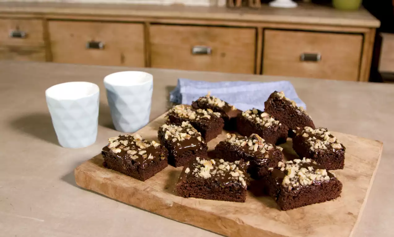 Brownies carrés au chocolat noir moelleux, parsemés de morceaux de noix dorés, disposés sur une planche en bois clair avec deux tasses blanches en relief et une serviette bleue en arrière-plan