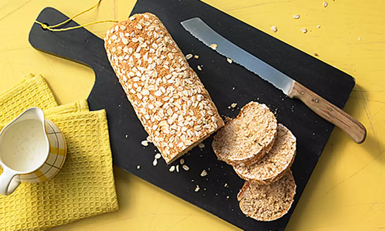 Hafer-Joghurt-Brot mit goldener Kruste und Haferflocken bestreut, auf schwarzem Holzbrett, daneben gelbes Geschirrtuch und passende Kanne