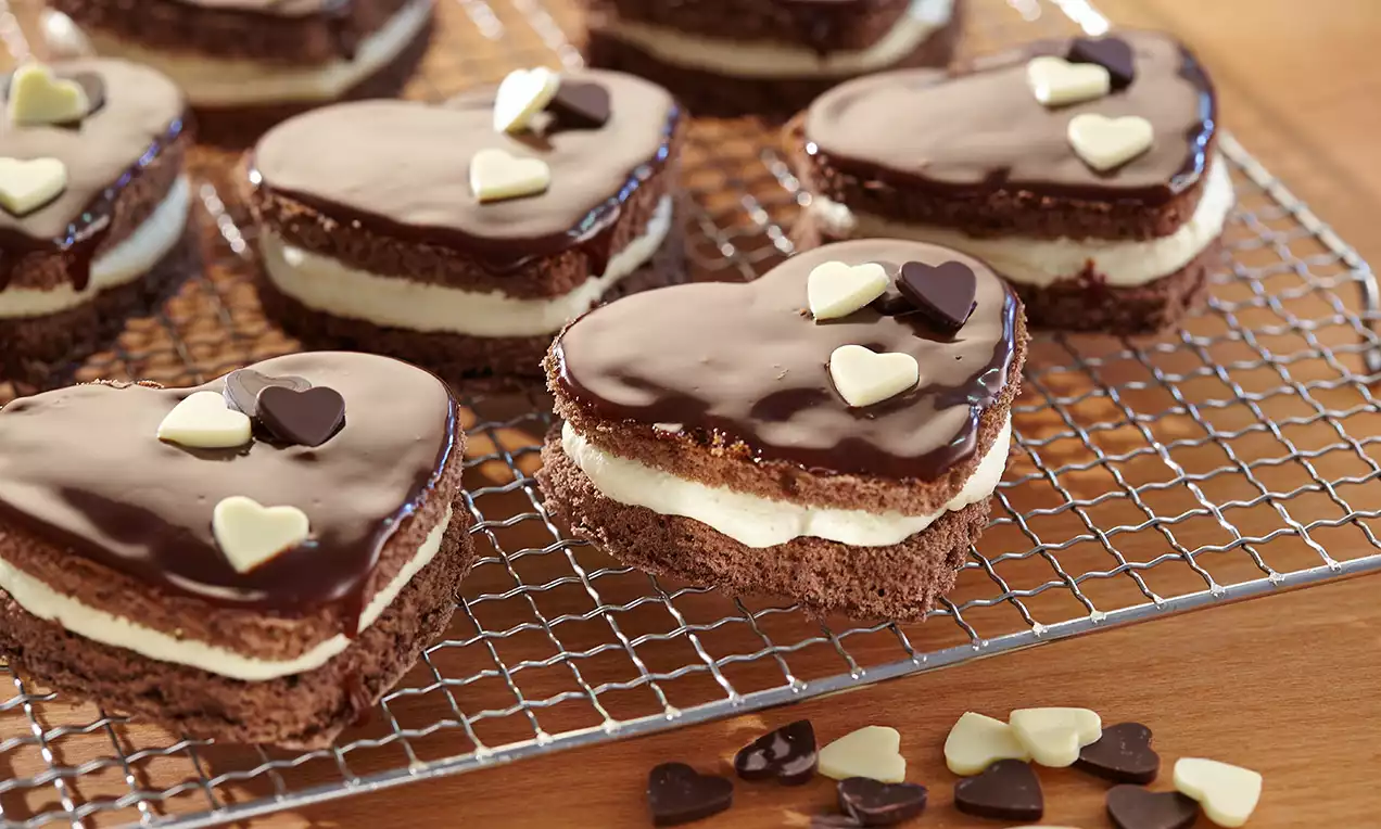 Herzförmige Schichtdesserts aus braunem Schokobiskuit mit heller Cremefüllung, glatter Schokoglasur und kleinen Herzstreuseln auf einem Gitter