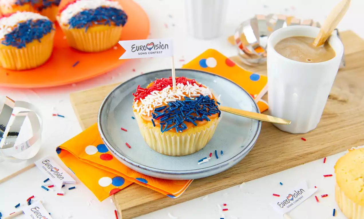 Vanillecupcake met rode, witte en blauwe sprinkles, versierd met een Eurovisievlaggetje, op een grijs bord met oranje servet en koffie ernaast