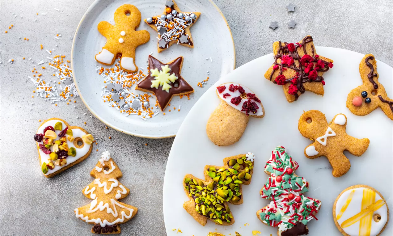 Коледни сладки с различни форми, глазура и цветни поръски, подредени върху бели чинии и декорирани с шоколад, ядки и сушени плодове