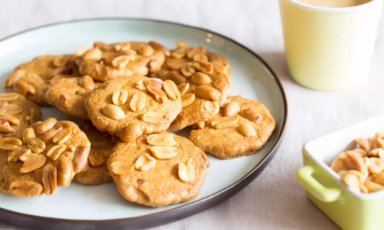 Ronde goudbruine pindakoekjes met een glanzende textuur en hele pinda's erop, gestapeld op een lichtblauw bord naast een kopje koffie