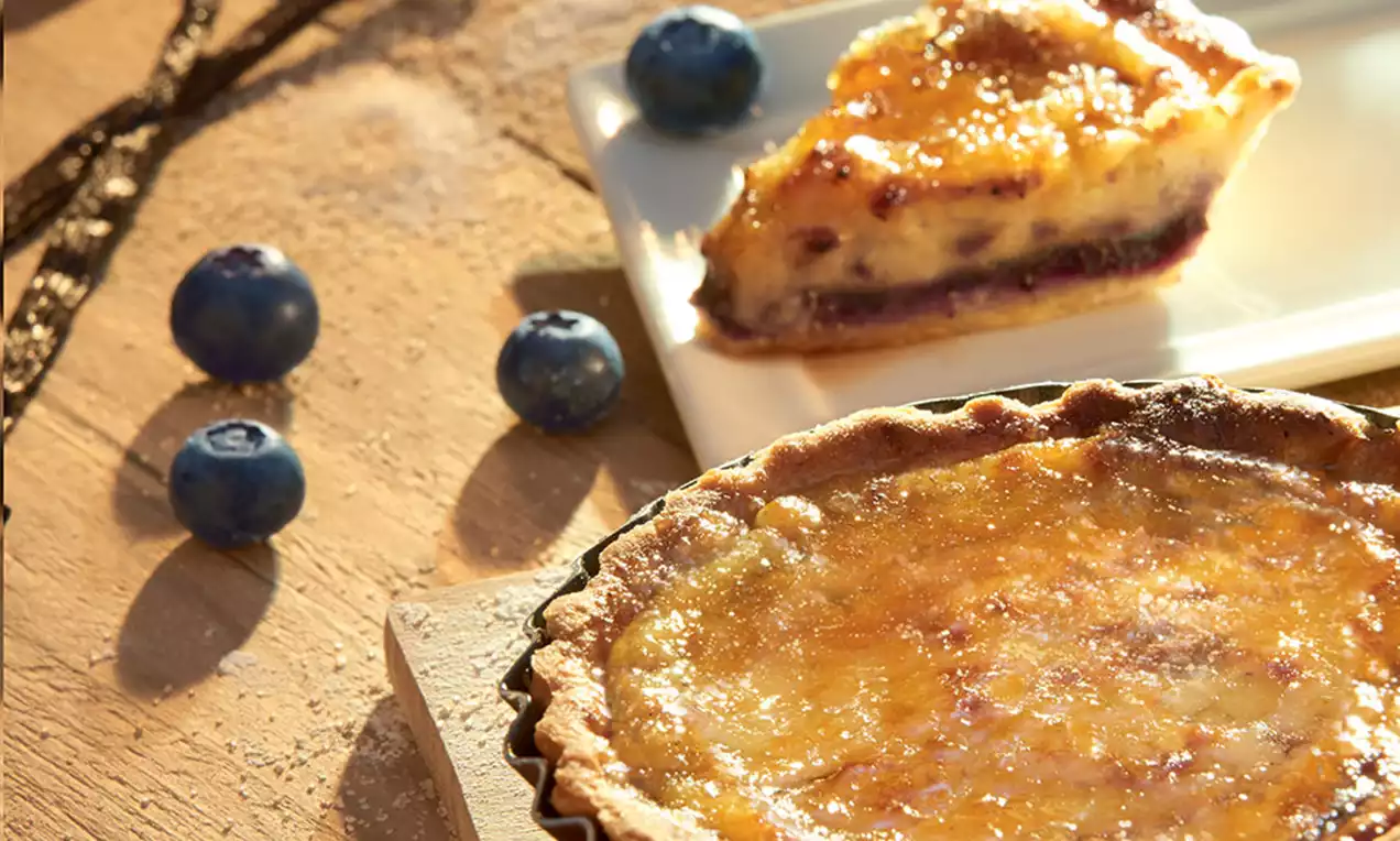 Crème brûlée-taartjes met goudbruin gekarameliseerde laag in ribbelvormige taartbodem, omgeven door verse blauwe bessen op een houten plank