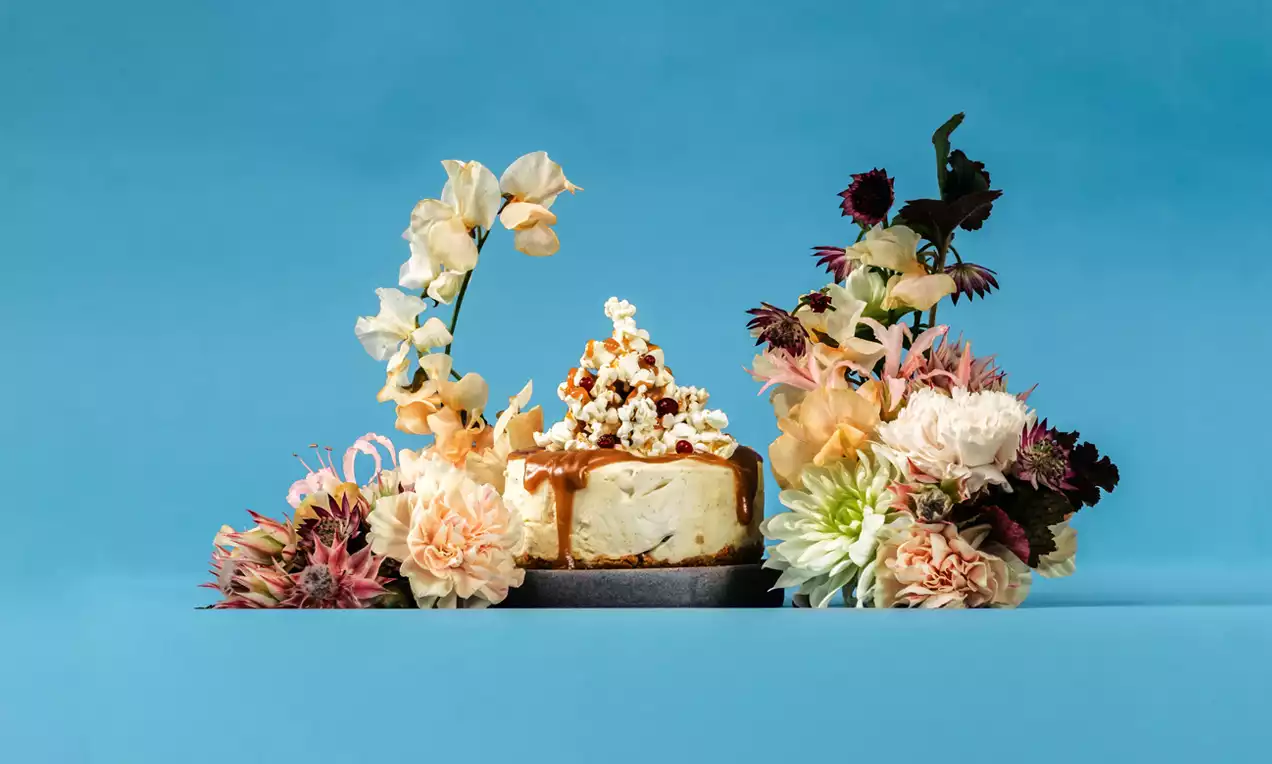 Kinuskilla valunut juustokakku pyöreällä tarjottimella, koristeltu kevyellä popcorniasettelulla ja ympäröity värikkäillä kukilla