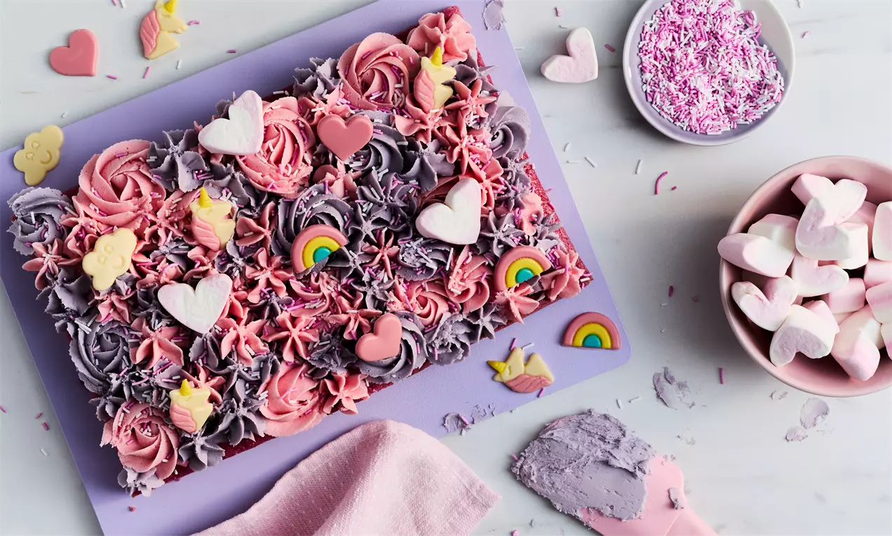Unicorn Magic Traybake with swirls of pink and purple frosting, rainbow and heart decorations on a pastel purple board