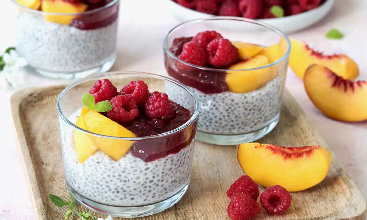 Chia-Pudding in klarem Glas mit hellgrauer Textur, darauf rote Himbeeren, gelbe Pfirsichspalten und dunkle Fruchtsoße auf Holztablett