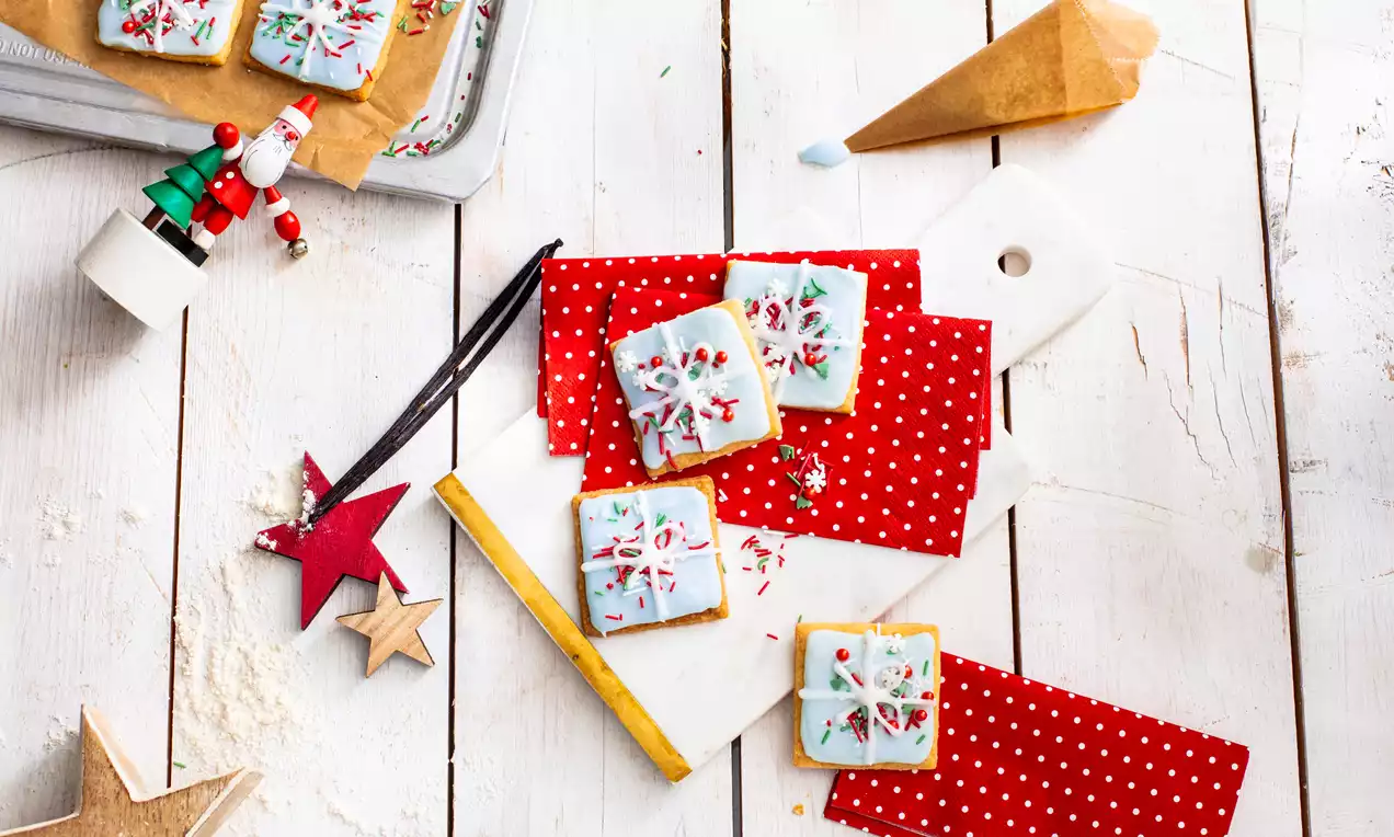 Fursecuri pătrate, ornate cu glazură albă decorată cu motive de iarnă colorate, aranjate pe șervețele roșii cu buline pe un fundal de lemn alb